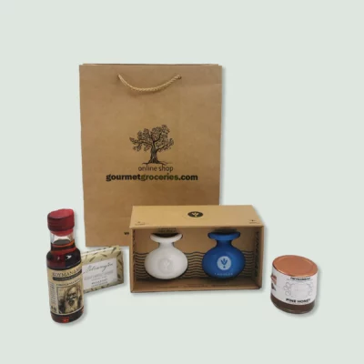 A brown paper gift bag with the logo and text "gourmet2.wearedoubledot.com" sits behind a selection of gourmet products. These include a small bottle of Commandaria red wine with a red cap, a bar of soap in green and white packaging, a box containing a white ceramic bottle of extra virgin olive oil and a blue ceramic bottle of balsamic vinegar, and a jar of pine honey.