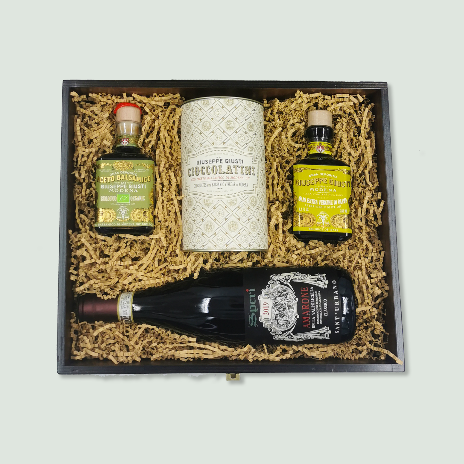 Box 20232 A A wooden gift box containing gourmet items arranged neatly in wood wool. The items include a bottle of balsamic vinegar, a tin of chocolate, a bottle of olive oil, and a bottle of red wine.
