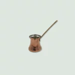 A traditional copper cezve with a long handle. The cezve has a hammered texture and is used for making Greek coffee.