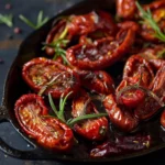A close-up of dried tomatoes on a plate, drizzled with olive oil and garnished with fresh herbs.
