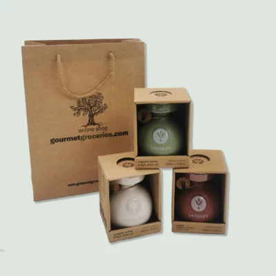 Three boxed ceramic bottles of Ladolea organic products displayed in front of a brown paper gift bag with a rope handle. The bag is branded with the logo "gourmet2.wearedoubledot.com." The bottles include organic extra virgin olive oil in white and green bottles, and organic balsamic vinegar with bergamot in a red bottle.
