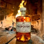 A glass of Spyropoulos Aged Tsipouro placed on a wooden table near a cozy fireplace, creating a warm and inviting atmosphere.