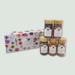 A box with five jars of mixed herbs by Vanissa, labeled "Lueur Florale." The jars have golden lids and contain colorful blends of edible flowers and herbs. The box features a floral design with various flowers illustrated on it, and the jars are arranged outside the box for display.